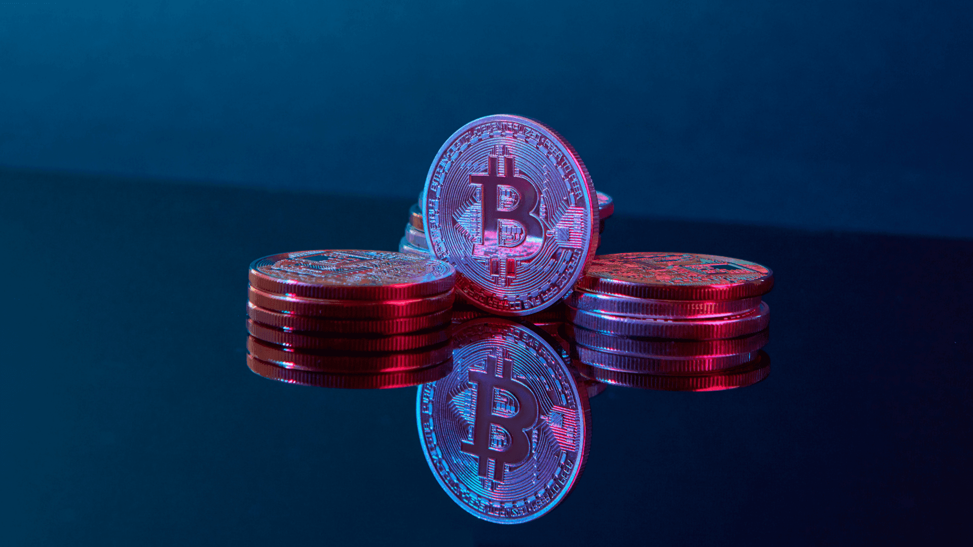 Physical Bitcoin tokens stacked on a reflective dark surface with vibrant blue and pink neon lighting.